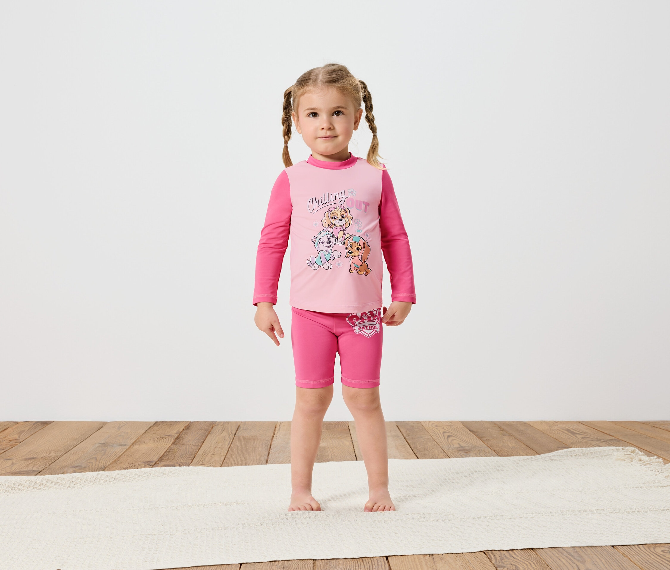 Malá holčička se zapletenými vlasy stojí bosá na krémovém koberci a má na sobě růžové tričko s dlouhým rukávem a šortky Tlapková patrola.