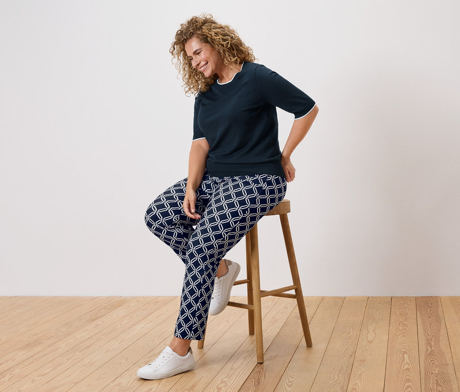 Žena s kudrnatými vlasy sedí na stoličce, oblečená v tmavě modré halence a vzorovaných kalhotách.