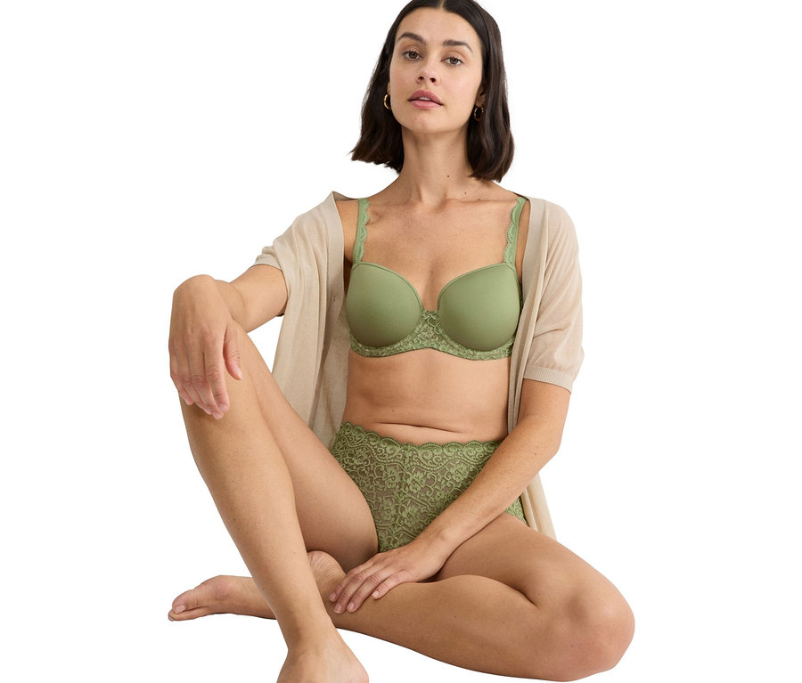 Žena sedí se zkříženýma nohama a má na sobě zelenou podprsenku a zelené krajkové kalhotky, spolu s béžovým svetrem.