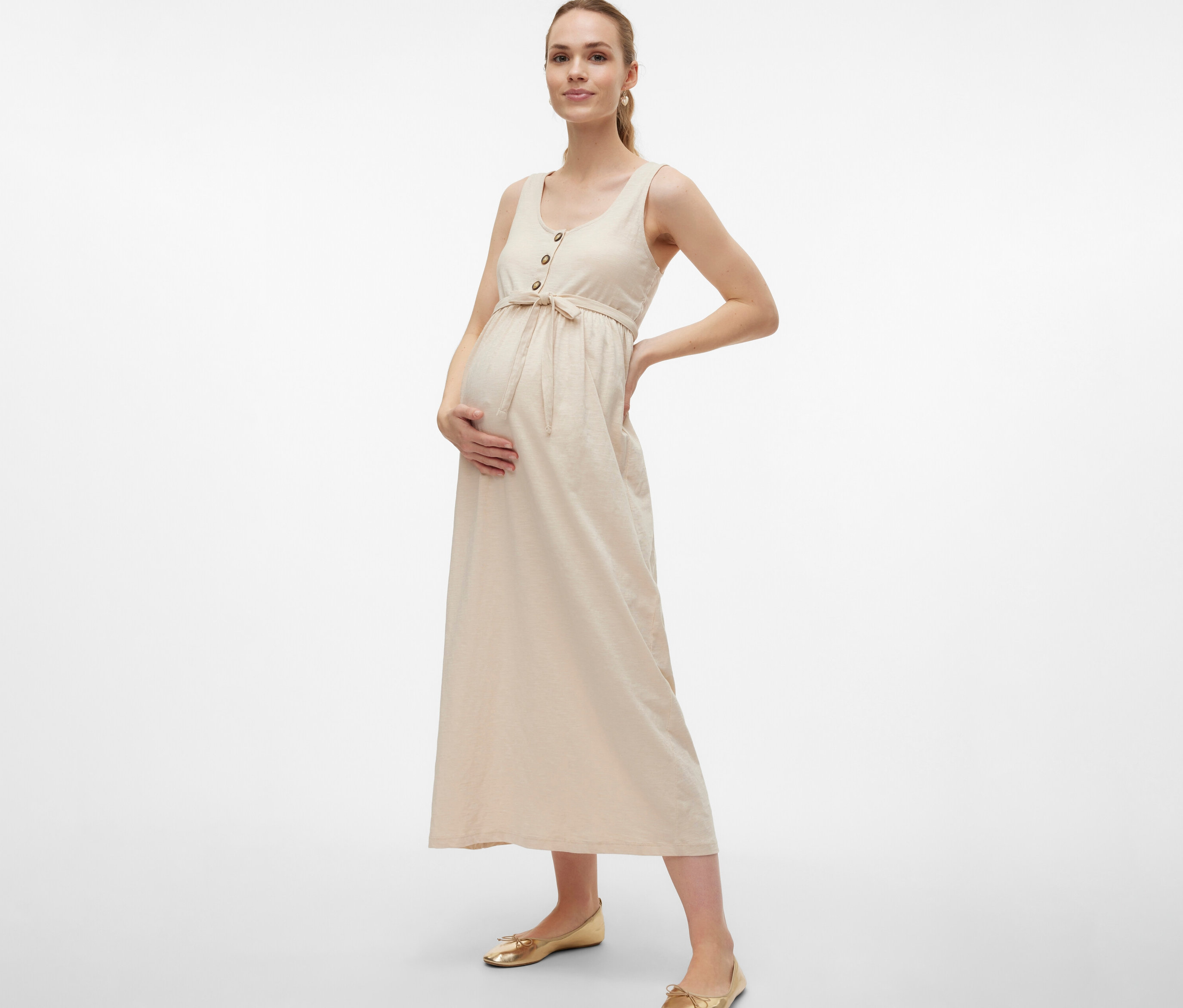 Těhotná žena v těhotenských šatech MAMALICIOUS »MLEVI LIA« s páskem a knoflíky pózuje s rukama na břiše a boku.