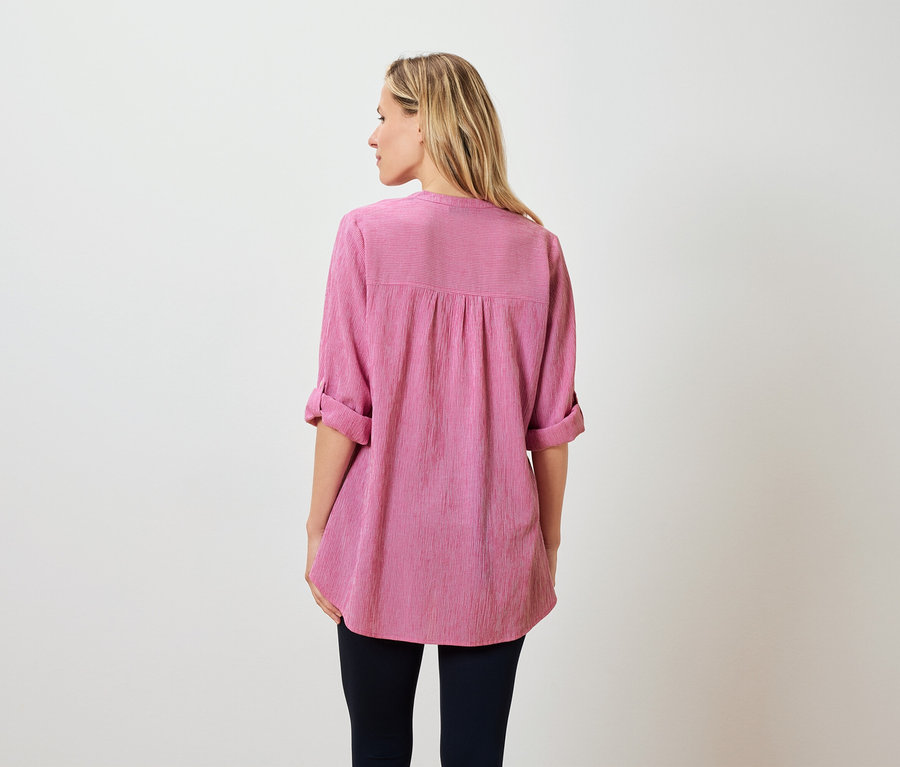 Pohled zezadu na ženu s blond vlasy po ramena, oblečenou v růžové halence a tmavých kalhotách.