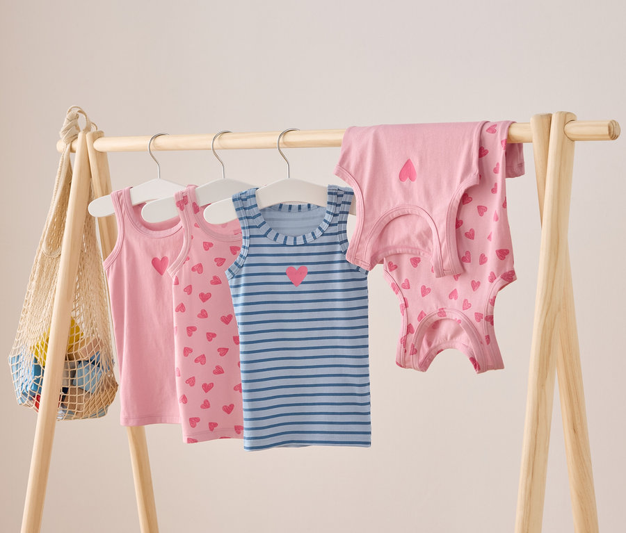 Na dřevěné tyči visí růžové a modré pruhované dětské tílka s motivem srdce.