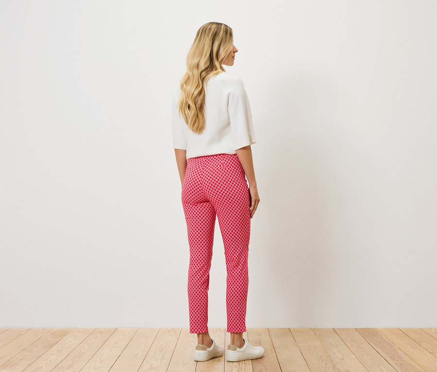 Pohled zezadu na ženu s blond vlnitými vlasy, oblečenou v bílé halence a červeno-růžových vzorovaných kalhotách a bílých botách.