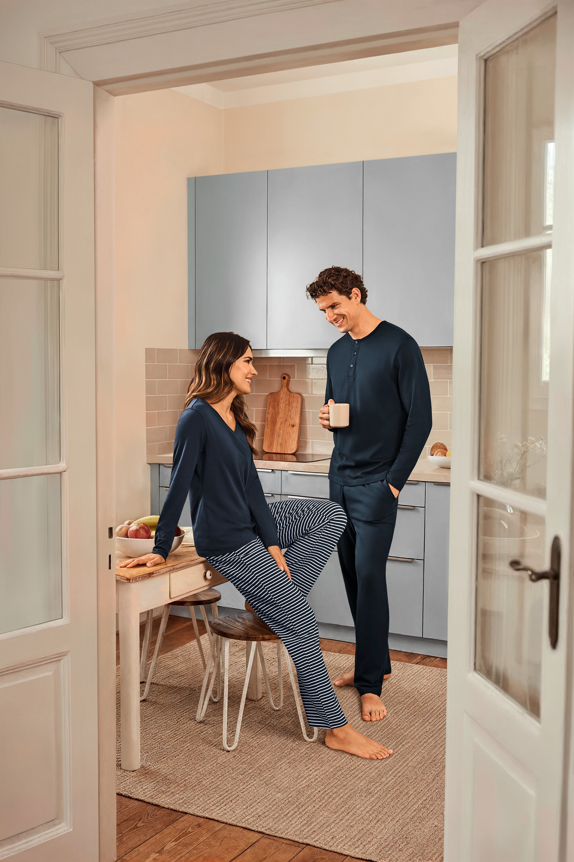 V kuchyni stojí žena sedící na stole v tmavě modrém pruhovaném pyžamu a tmavě modrém topu a muž, který drží šálek a má na sobě tmavě modré pyžamo.