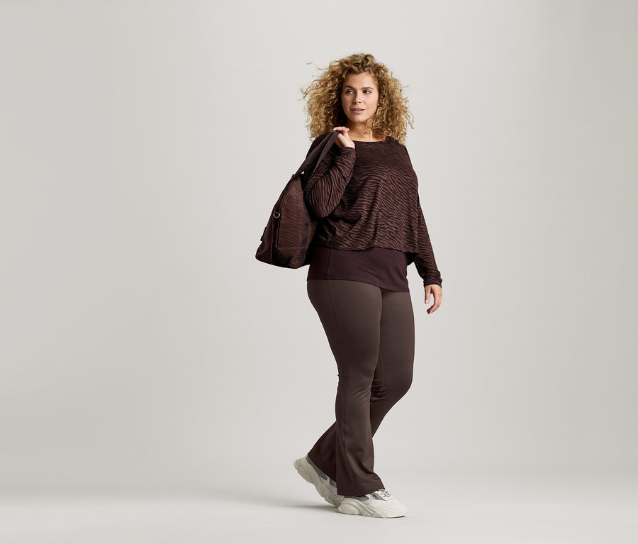 Žena s kudrnatými vlasy má na sobě hnědý top se zvířecím potiskem, hnědé legíny a tenisky. Přes rameno má hnědou tašku.