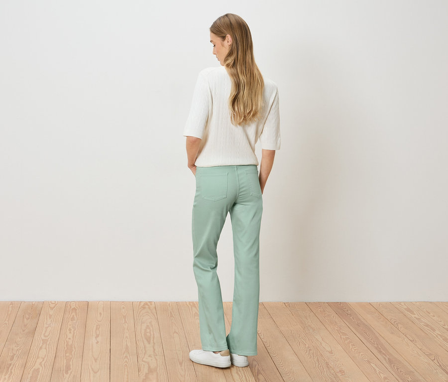 Pohled zezadu na ženu s blond vlasy v bílém svetru a světle zelených kalhotách.