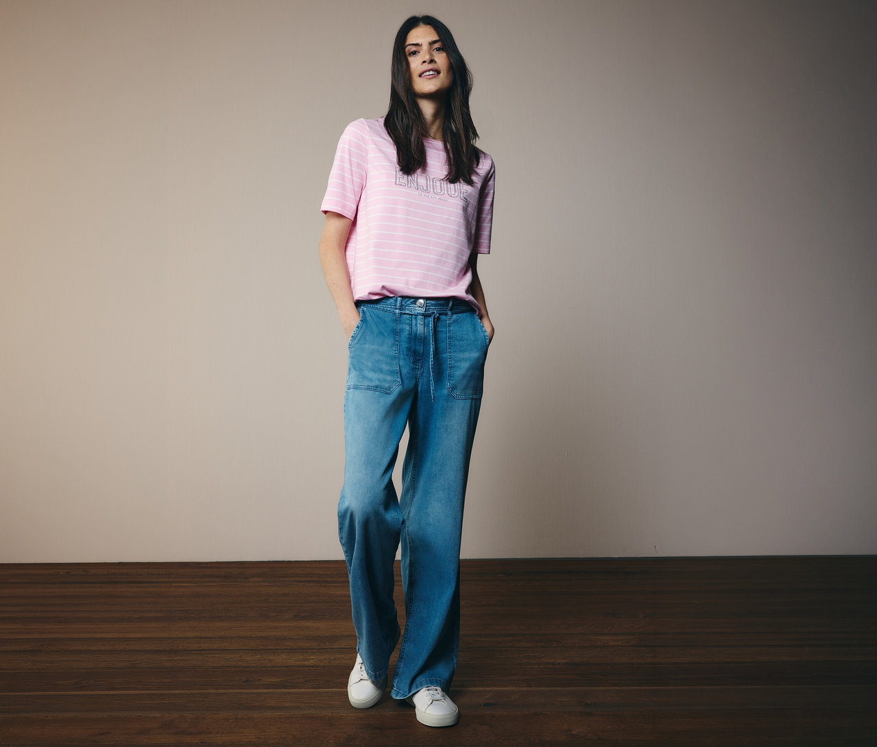 Žena v tričku CECIL s růžovými pruhy a širokých džínách a bílých teniskách pózuje s rukama v kapsách.