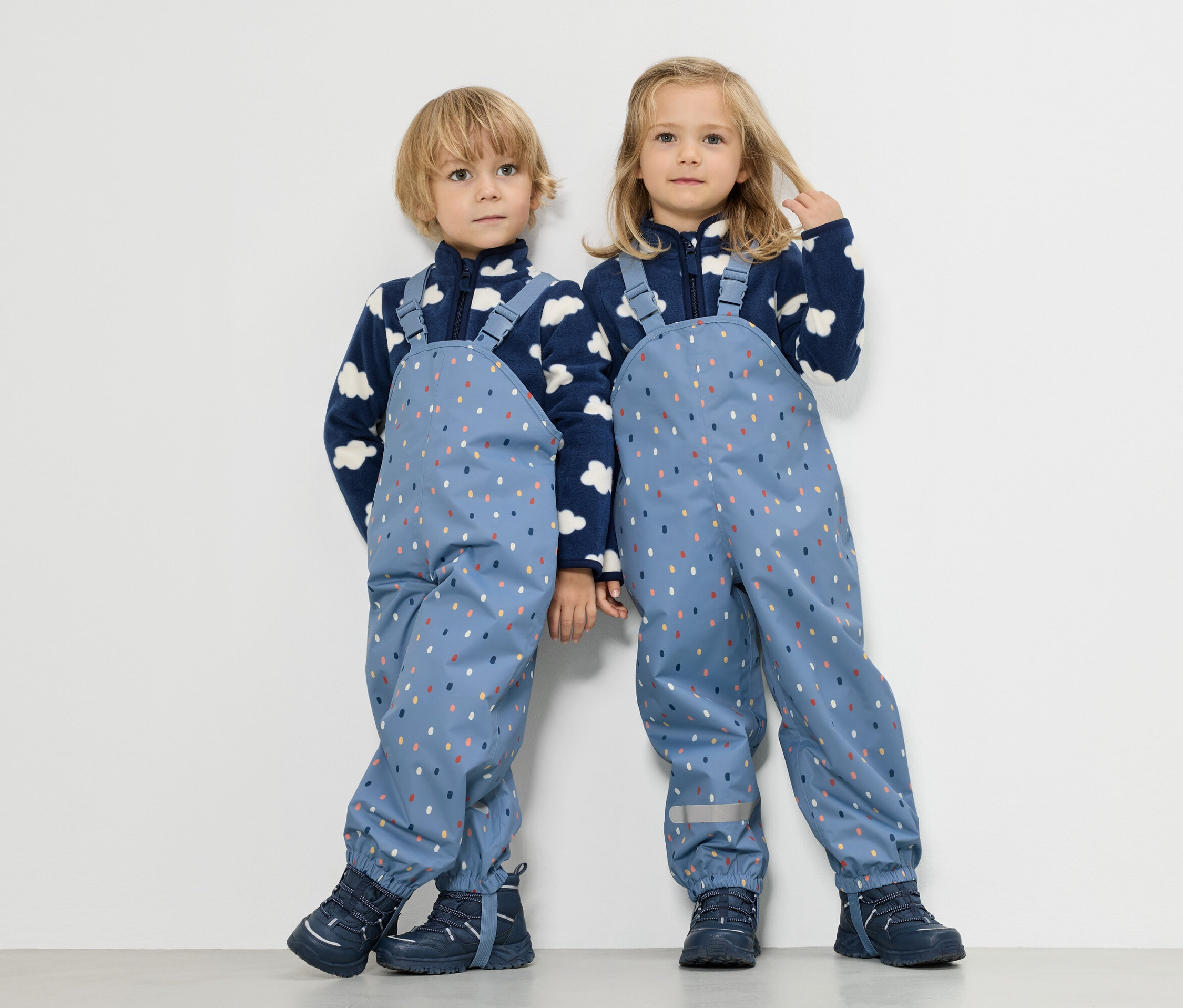 Dvě děti stojí vedle sebe, chlapec a dívka, oba mají na sobě modré fleecové bundy s motivem mraků a modré nepromokavé kalhoty s barevnými tečkami.