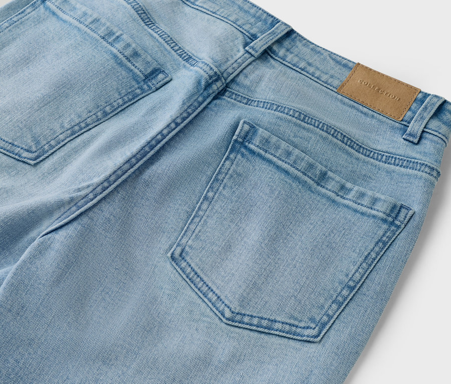 Detailní záběr na zadní část světle modrých džínů s kapsami a hnědým štítkem 'Collection'.