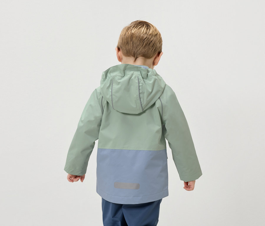 Pohled zezadu na dítě s blond vlasy v dvoubarevné bundě zelené a modré barvy.