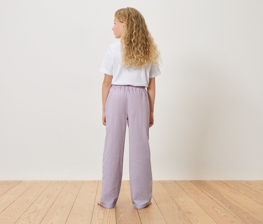 Pohled zezadu na mladou dívku s blond kudrnatými vlasy, oblečenou v bílém tričku a světle fialových pruhovaných kalhotách.