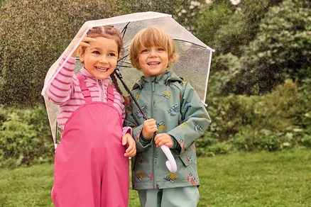 Dvě smějící se děti, dívka v růžových lacláčích a chlapec v zelené bundě s motivem bagru, stojí v dešti pod průhledným deštníkem.