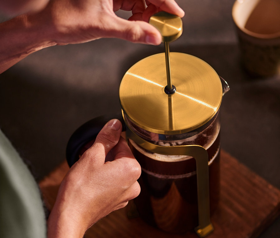Osoba stlačuje zlatou konvičku french press 800 ml na 6 šálků na dřevěném podkladu.