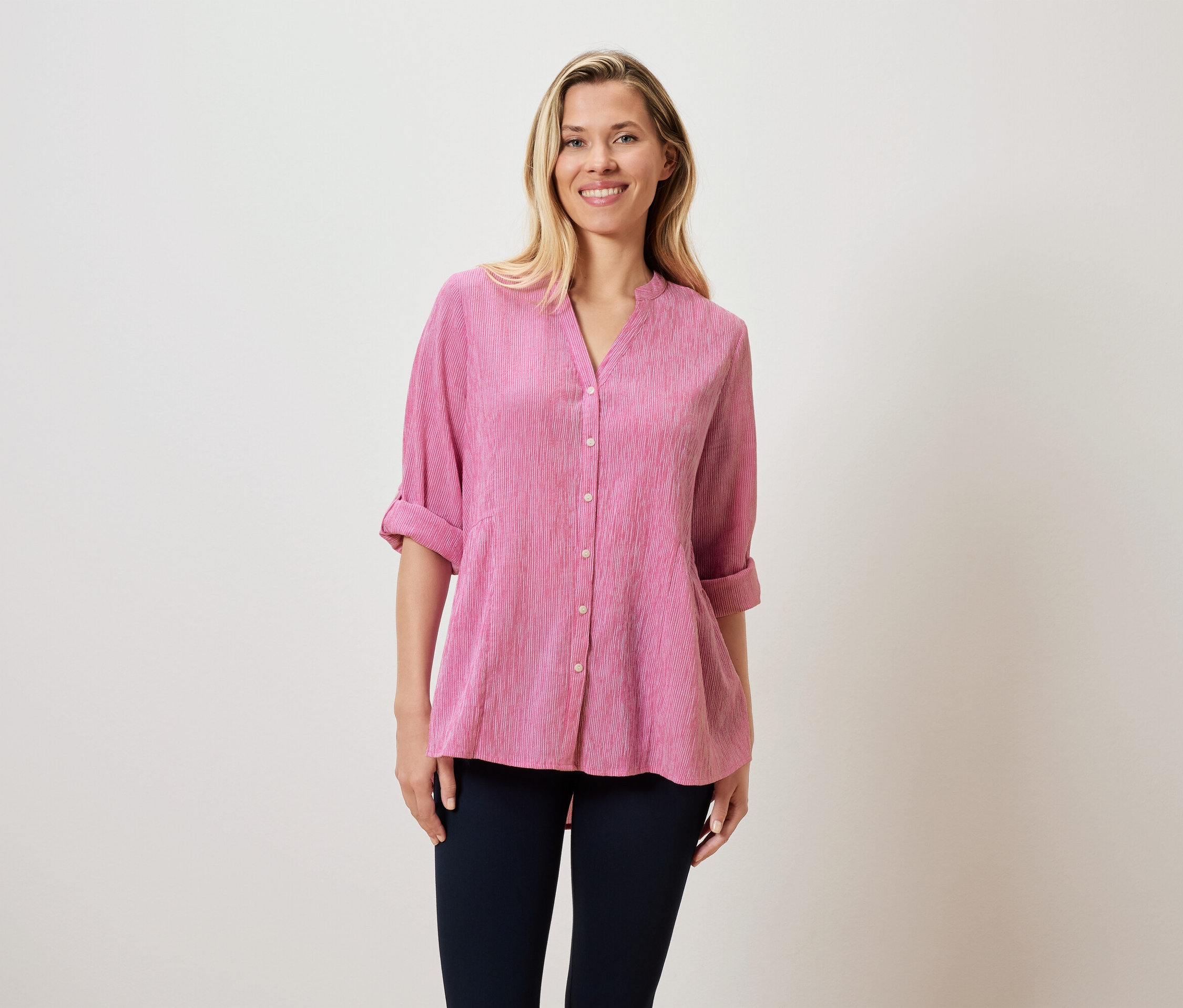 Žena s blond vlasy má na sobě růžovou blůzu a tmavé kalhoty.