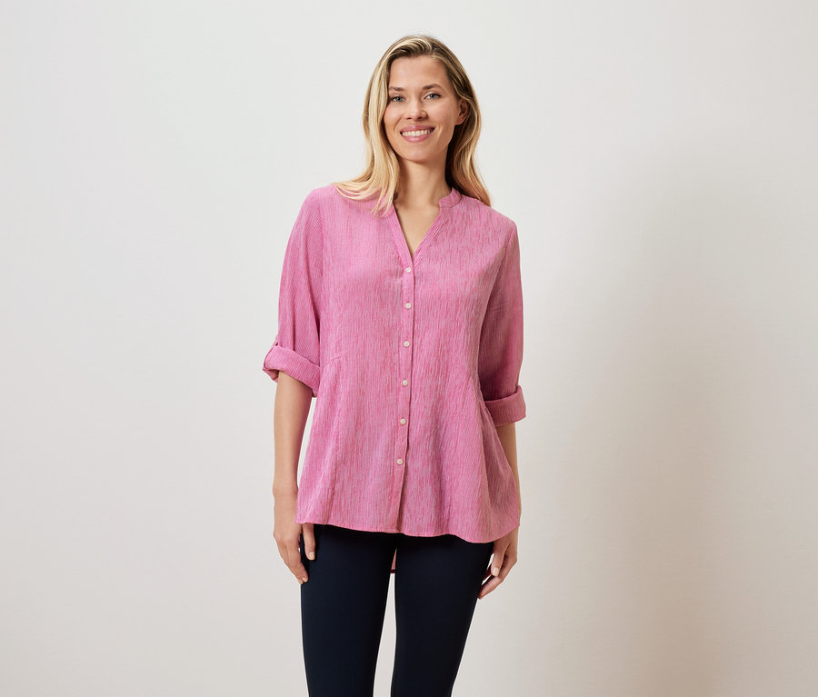 Žena s blond vlasy má na sobě růžovou blůzu a tmavé kalhoty.