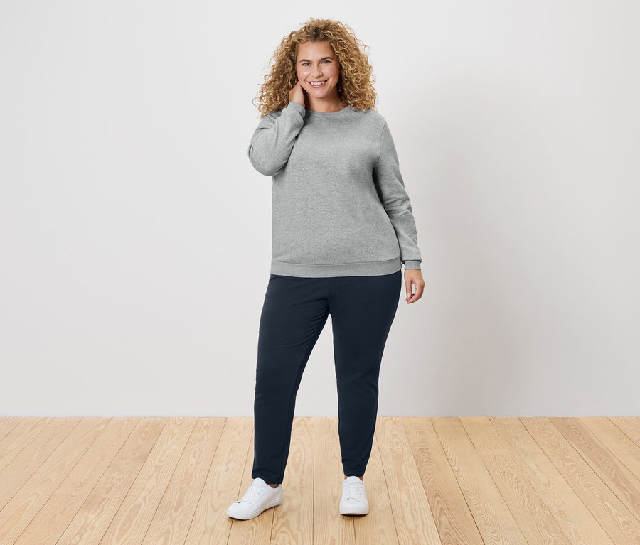 Žena s kudrnatými vlasy v šedém svetru a tmavě modrých kalhotách.