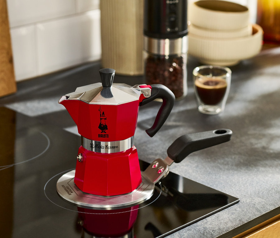 Červený kávovar na espresso stojí na varné desce vedle šálku espressa.