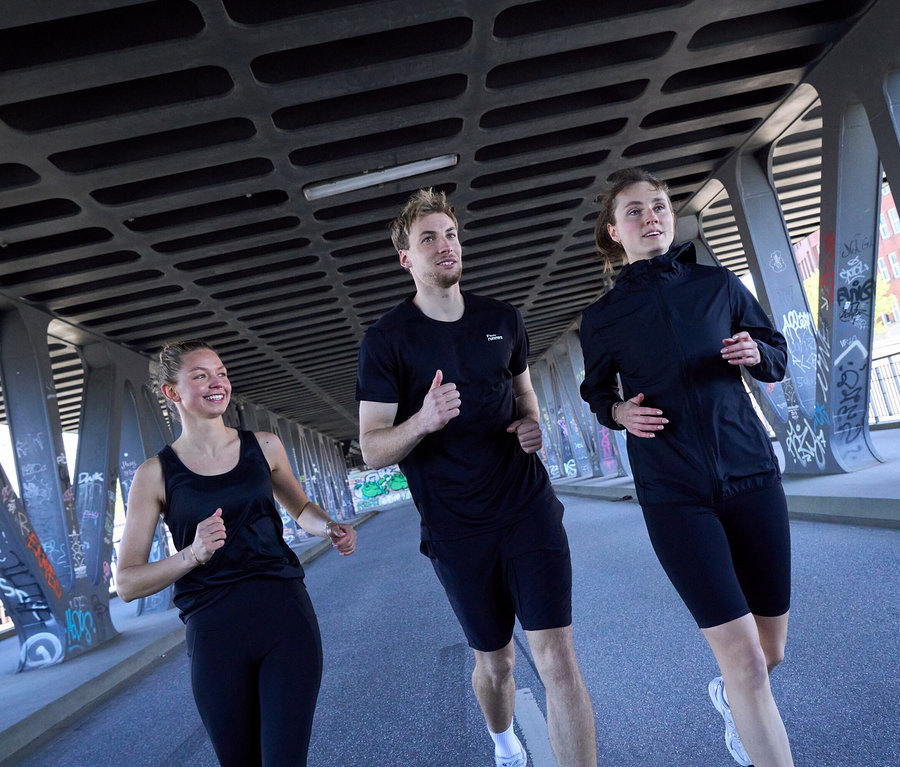 Tři lidé běží pod mostem: Žena v černém sportovním topu a černých sportovních legínách »ActivePower«, muž v černém sportovním tričku a černých sportovních šortkách, a žena v černé větrovce »packable« a černých sportovních cyklistických šortkách »ActivePower«.