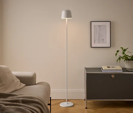 Moderní obývací pokoj s béžovou pohovkou, bílou stojací lampou a tmavě šedou skříňkou. Osvětlená lampa stojí mezi pohovkou a skříňkou, na které jsou knihy a pokojová rostlina pod zarámovaným obrázkem.
