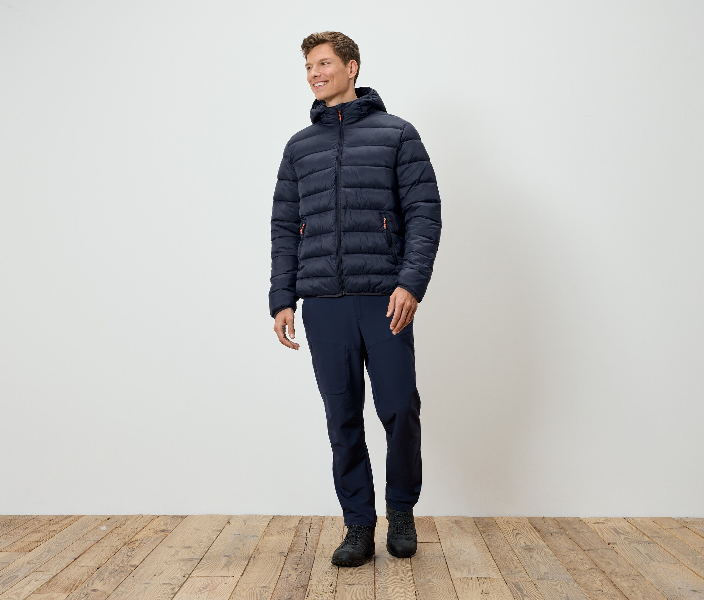 Muž stojí a má na sobě tmavě modrou prošívanou bundu, tmavě modré kalhoty a černé boty.