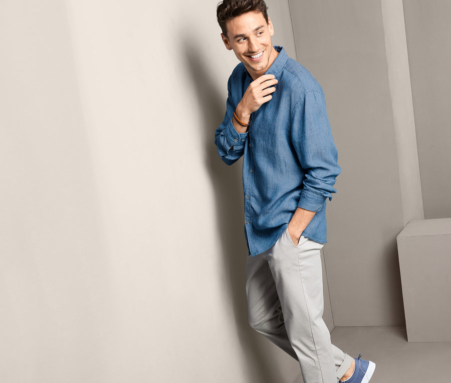 Muž se usmívá a opírá se o zeď v modré košili a šedých kalhotách.