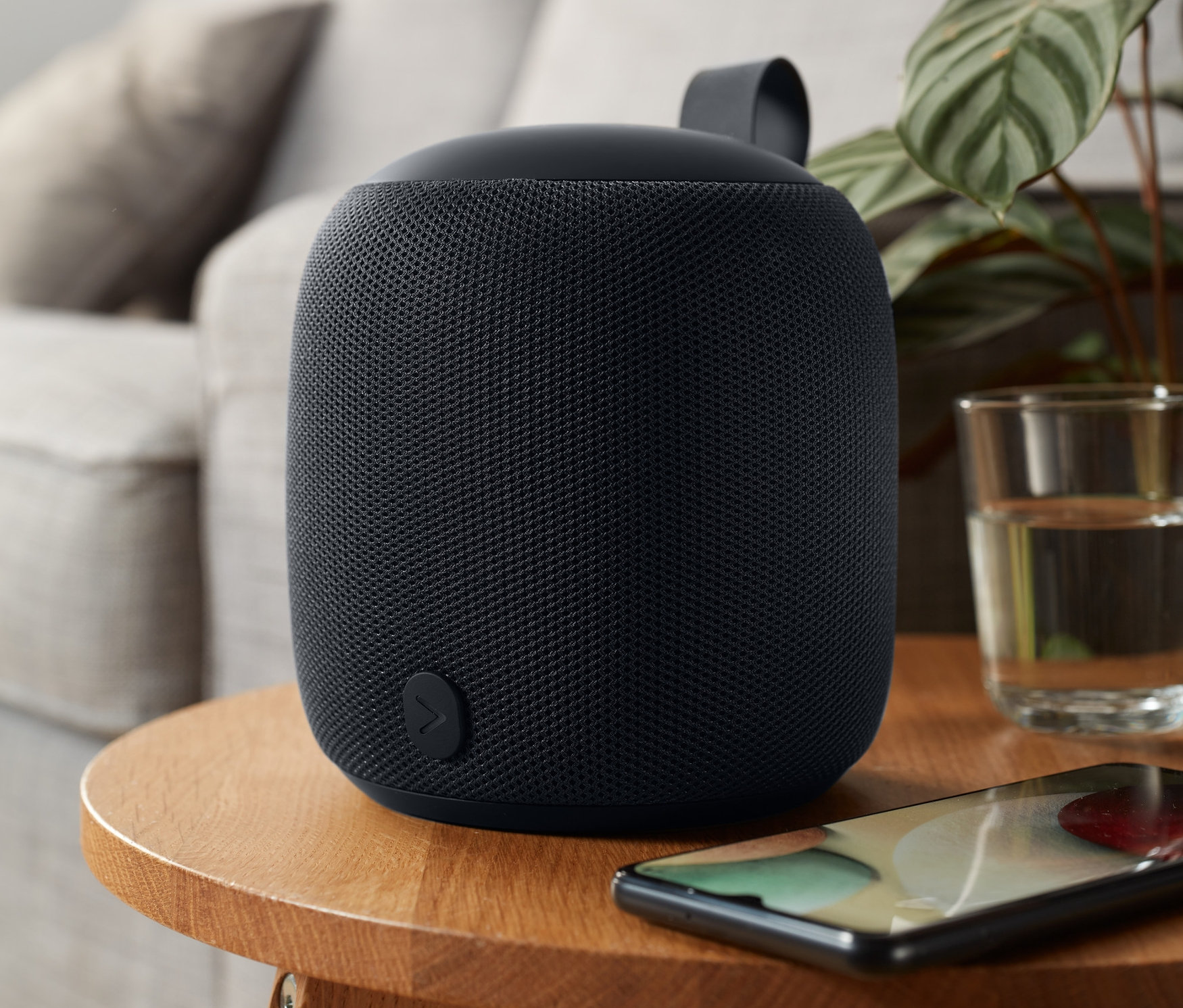 Antracitový designový reproduktor Bluetooth®, M, umístěný na dřevěném stolku vedle sklenice s vodou a mobilního telefonu.