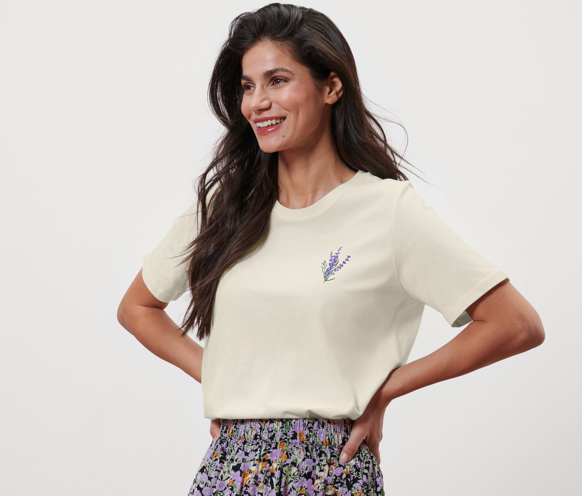Žena pózuje v krémovém tričku s potiskem levandule a květinovou sukni.