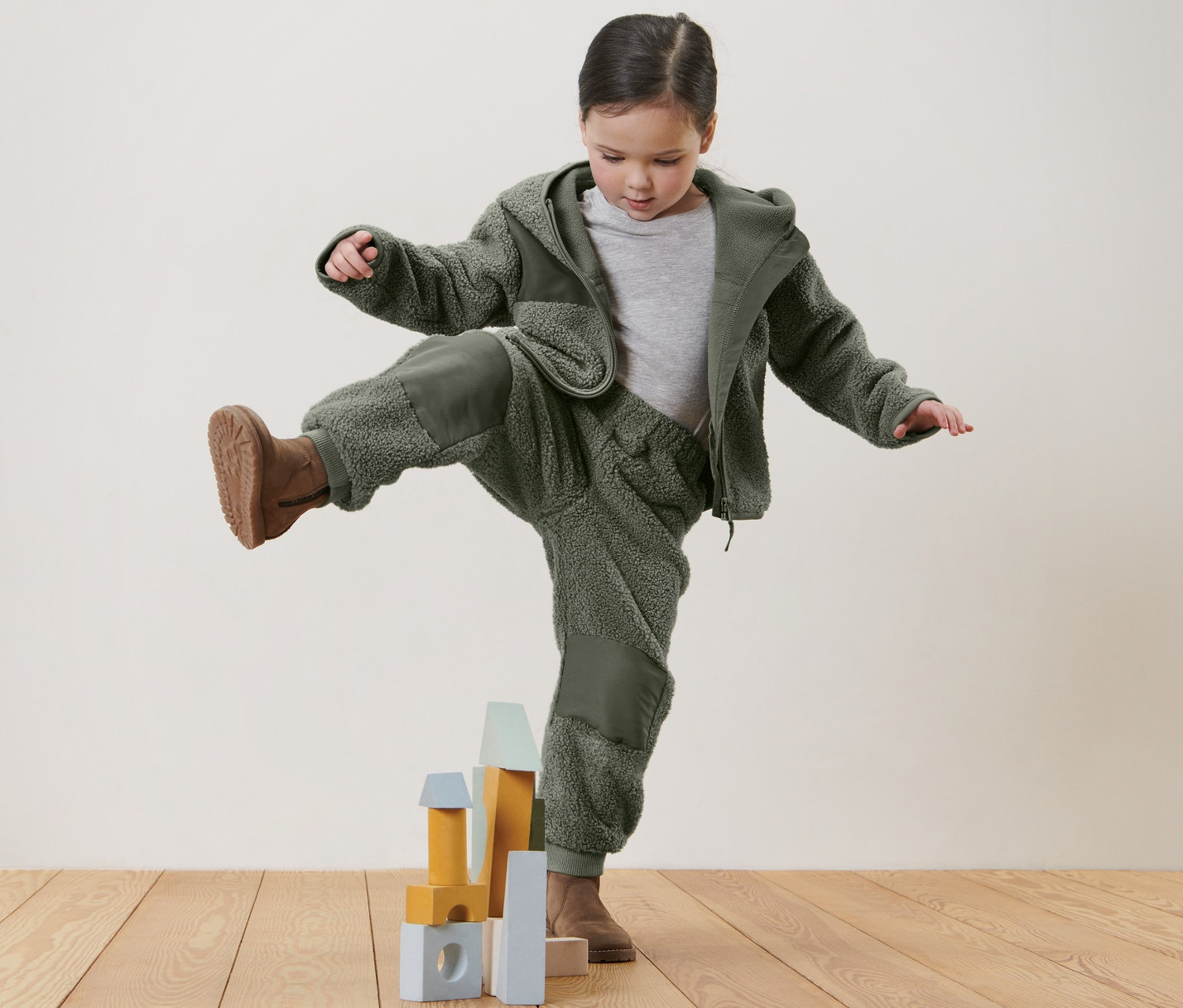 Dívka stojí na jedné noze na dřevěné podlaze a hraje si s dětskými plyšovými kalhotami a kostkami.