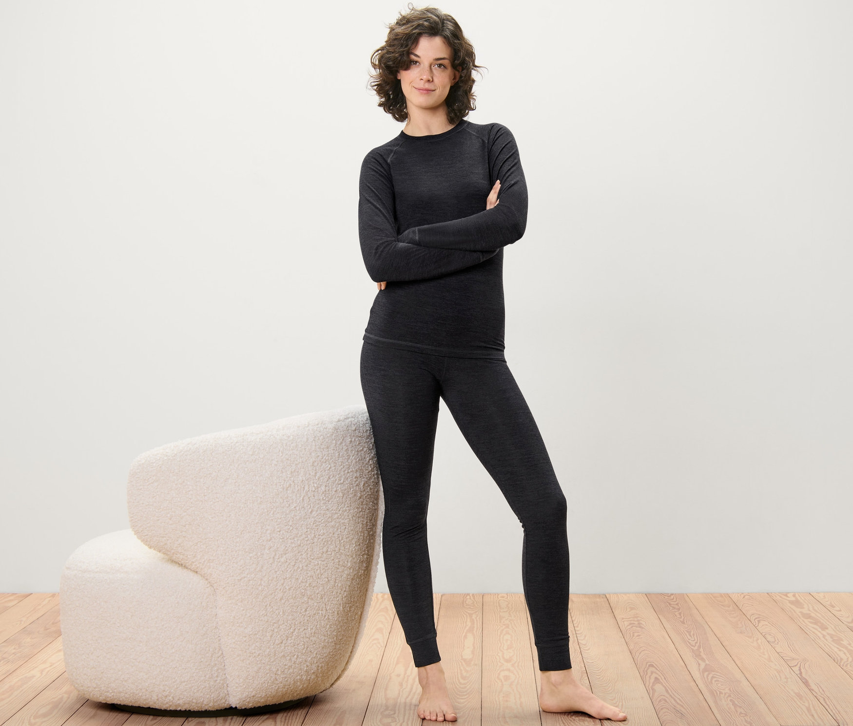 Žena pózuje v dlouhých spodkách s vlnou s certifikací RWS a horním dílu na dřevěné podlaze vedle krémového křesla.