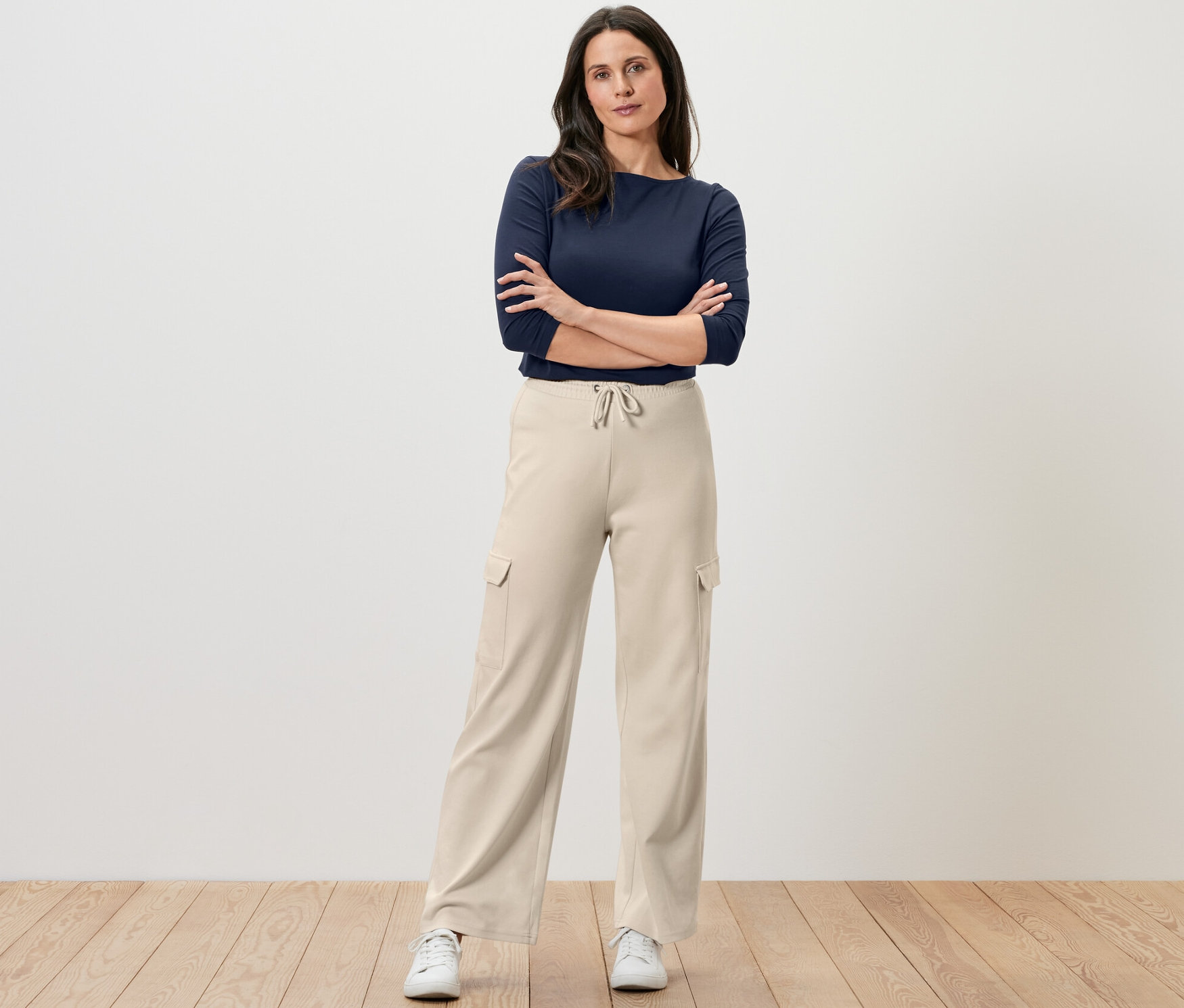 Žena stojí s rukama zkříženýma na prsou a má na sobě krémově bílé teplákové kalhoty s cargo kapsami a tmavě modrý top.