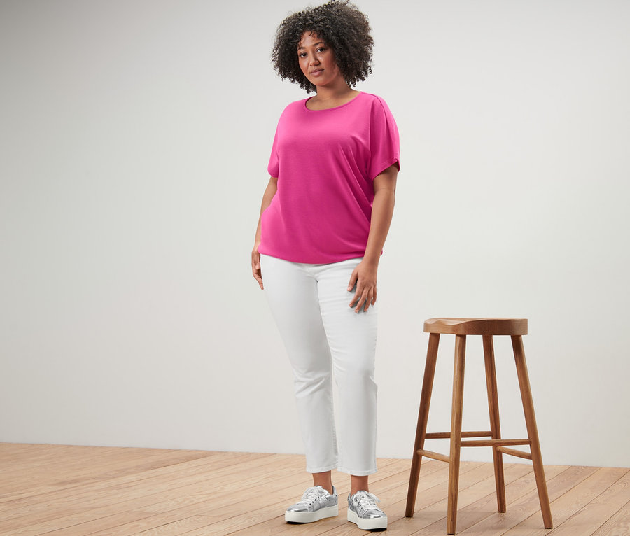 Žena stojí vedle dřevěné stoličky a má na sobě halenkové tričko a krémové džíny colored – Fit »Emma«.