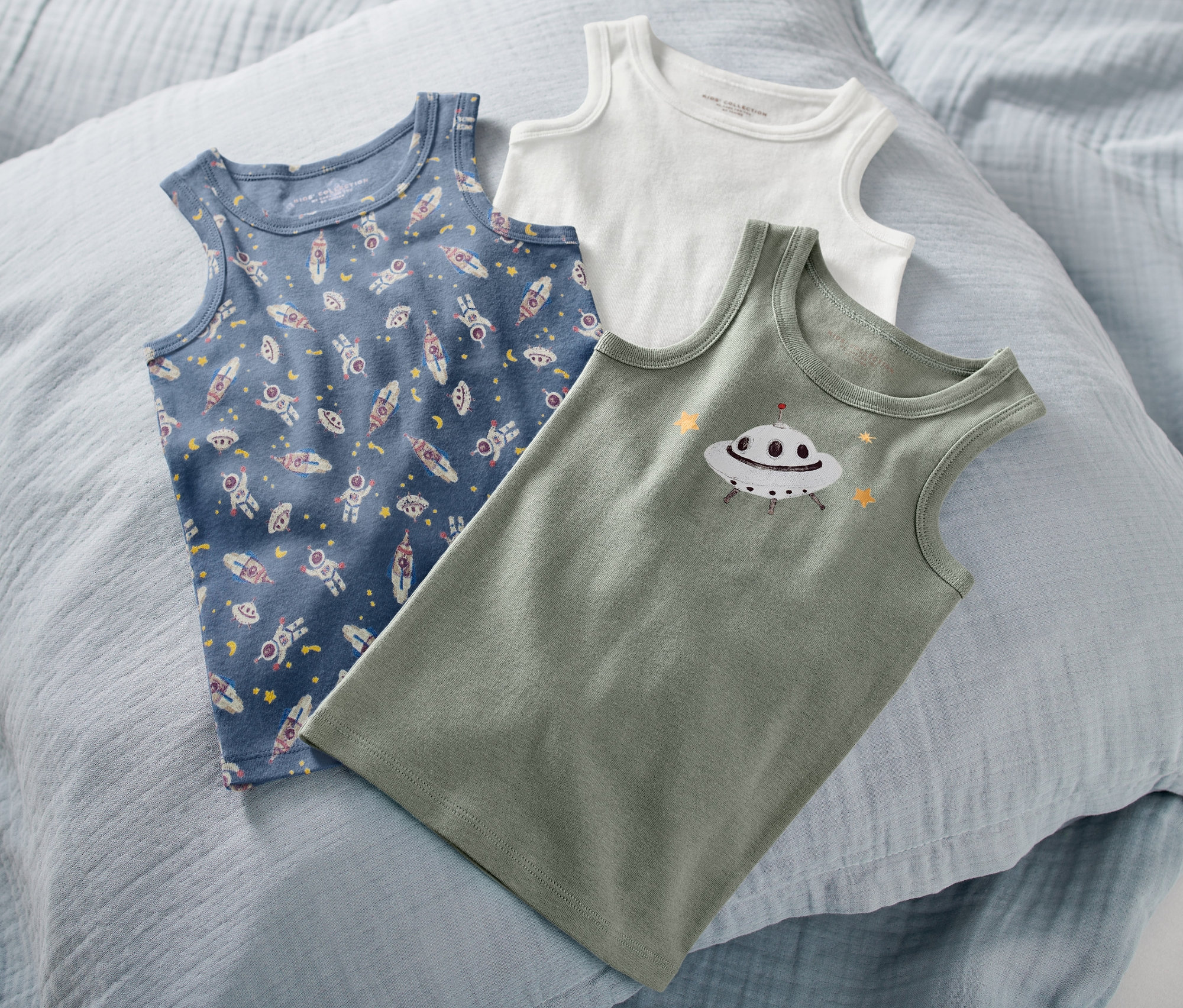 Na světle modrém polštáři leží tři dětská tílka. Jedno modré s potiskem vesmírných lodí, zelené s potiskem ufa a bílé.
