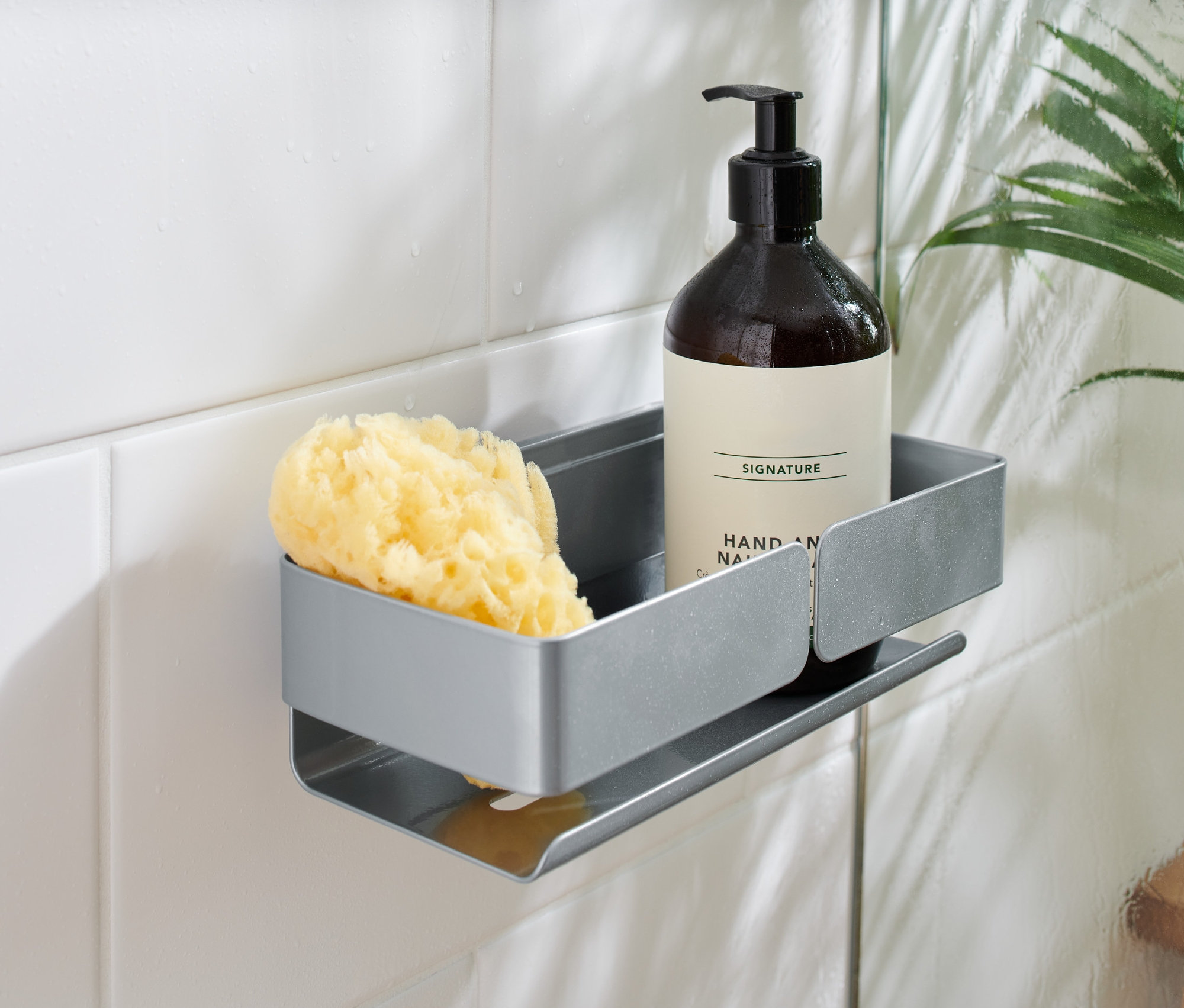 Šedá polička do sprchy připevněná na bílých kachličkách drží hnědou lahvičku s pumpičkou a žlutou houbu.