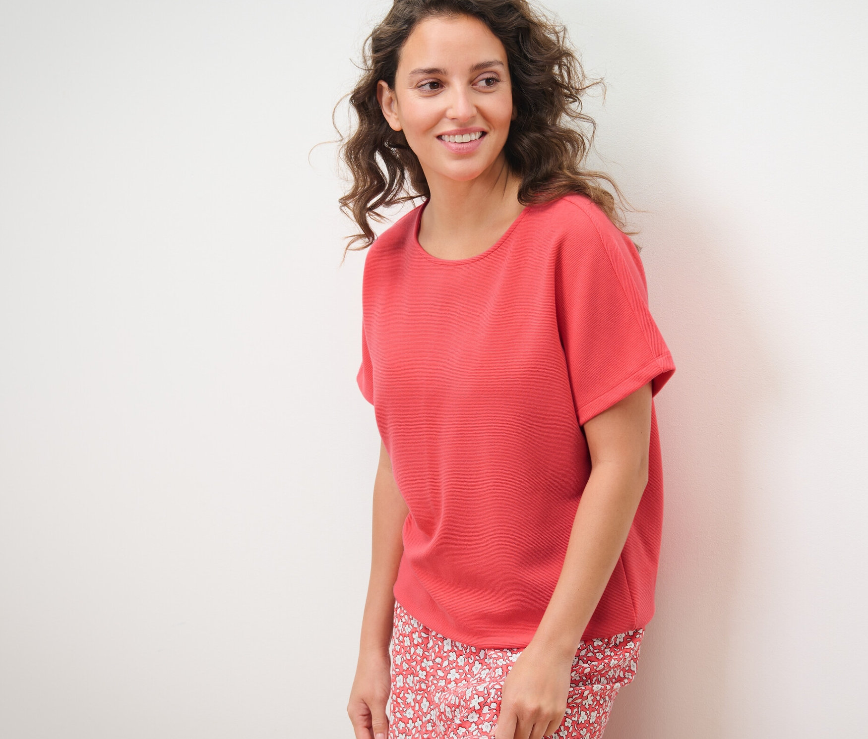 Žena pózuje na bílém pozadí a má na sobě korálové halenkové tričko.