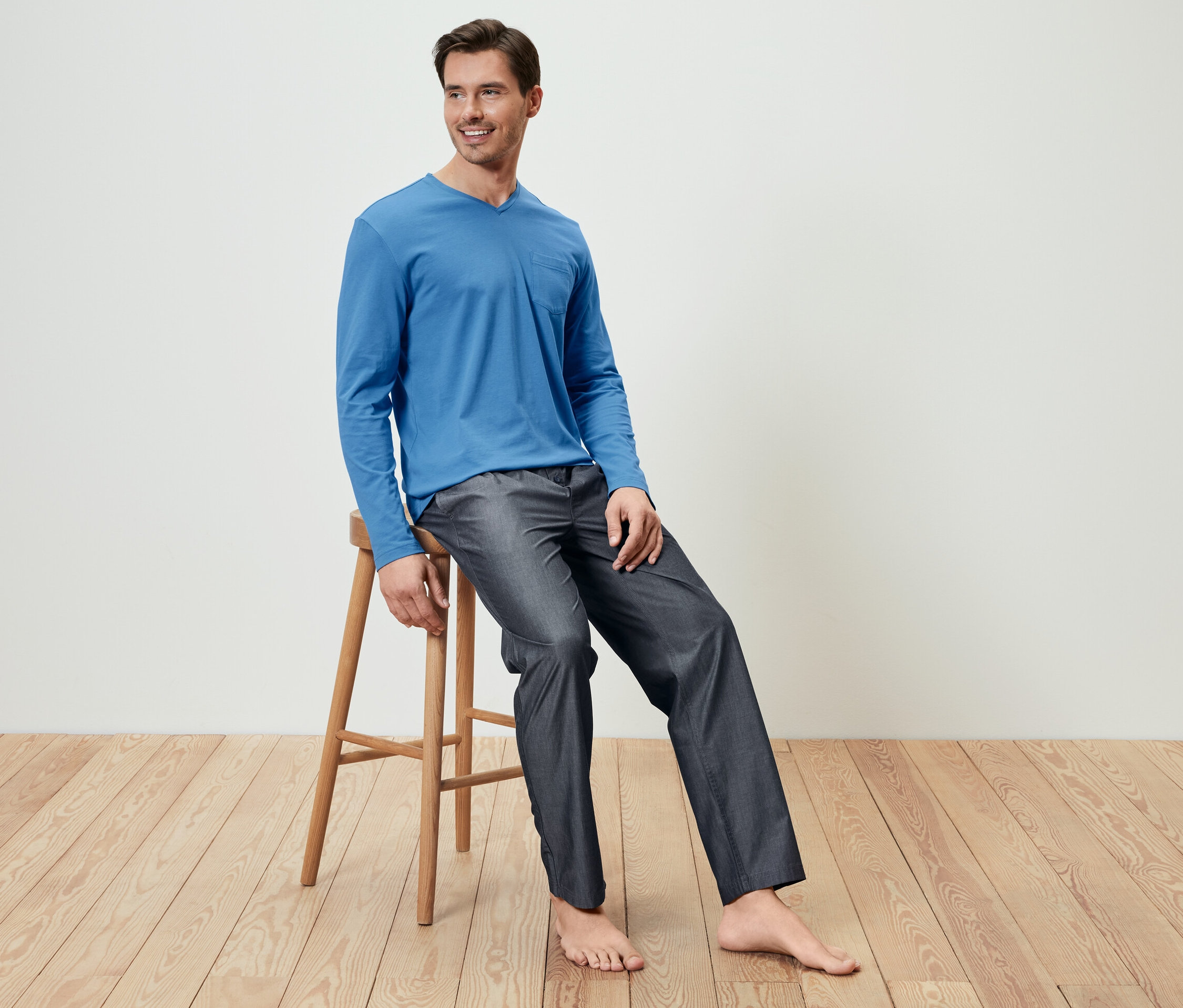 Muž sedí na dřevěné stoličce a má na sobě modré pyžamové tričko a tkané kalhoty.