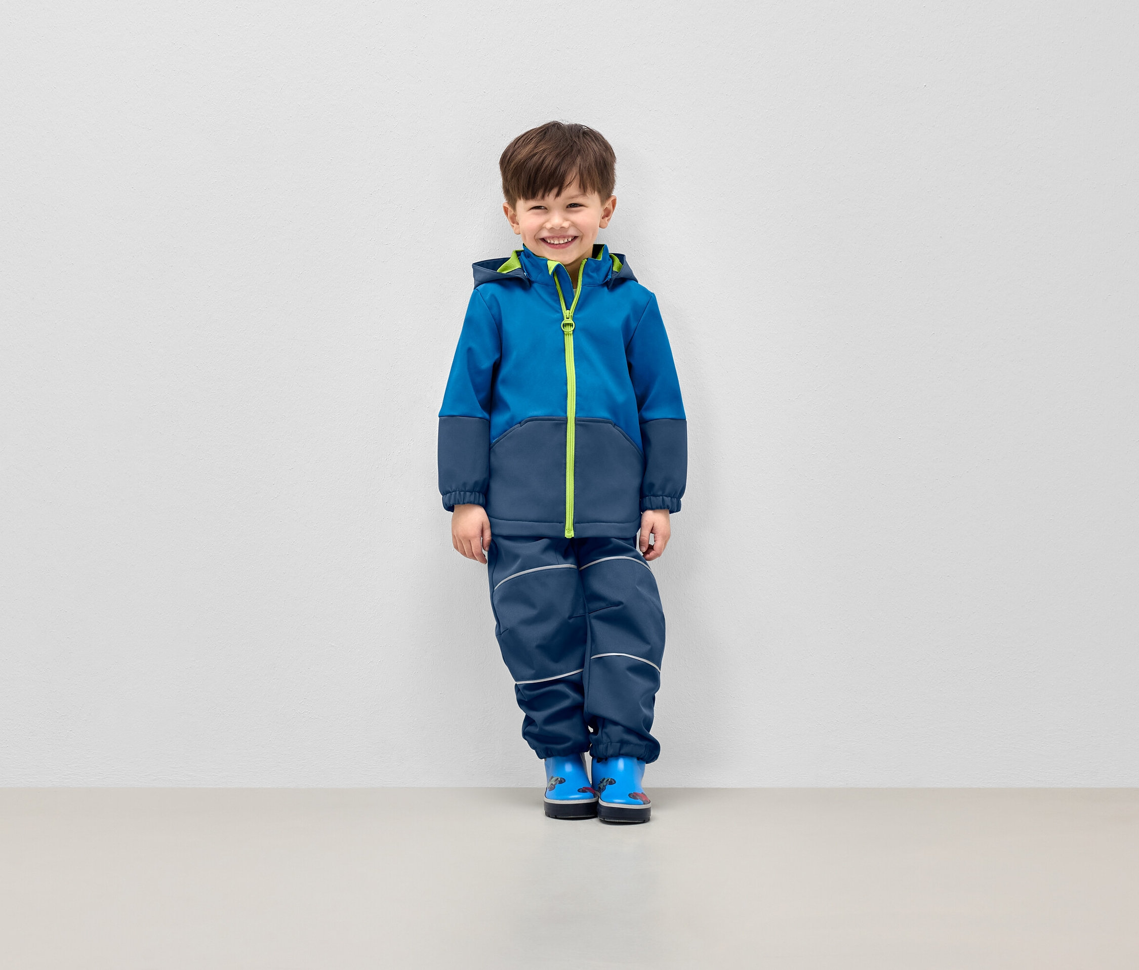 Usmívající se chlapec pózuje v modré bundě s kapucí a tmavě modrých kalhotách s reflexními pruhy.