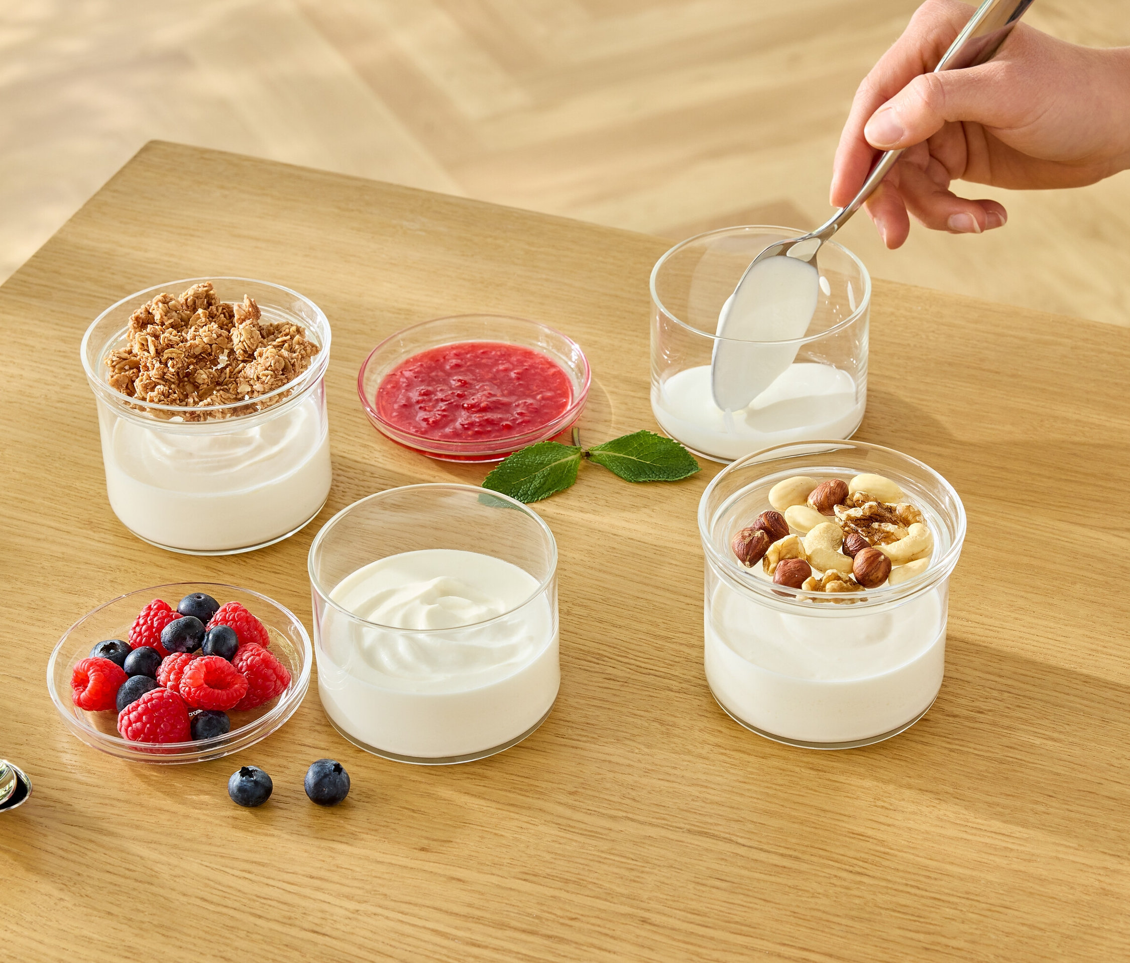 Ruka lžící nabírá bílý jogurt do sklenice. Na stole jsou další sklenice s jogurtem, müsli, ovocem a ořechy.