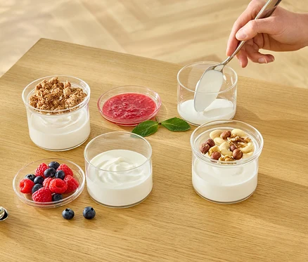 Ruka lžící nabírá bílý jogurt do sklenice. Na stole jsou další sklenice s jogurtem, müsli, ovocem a ořechy.