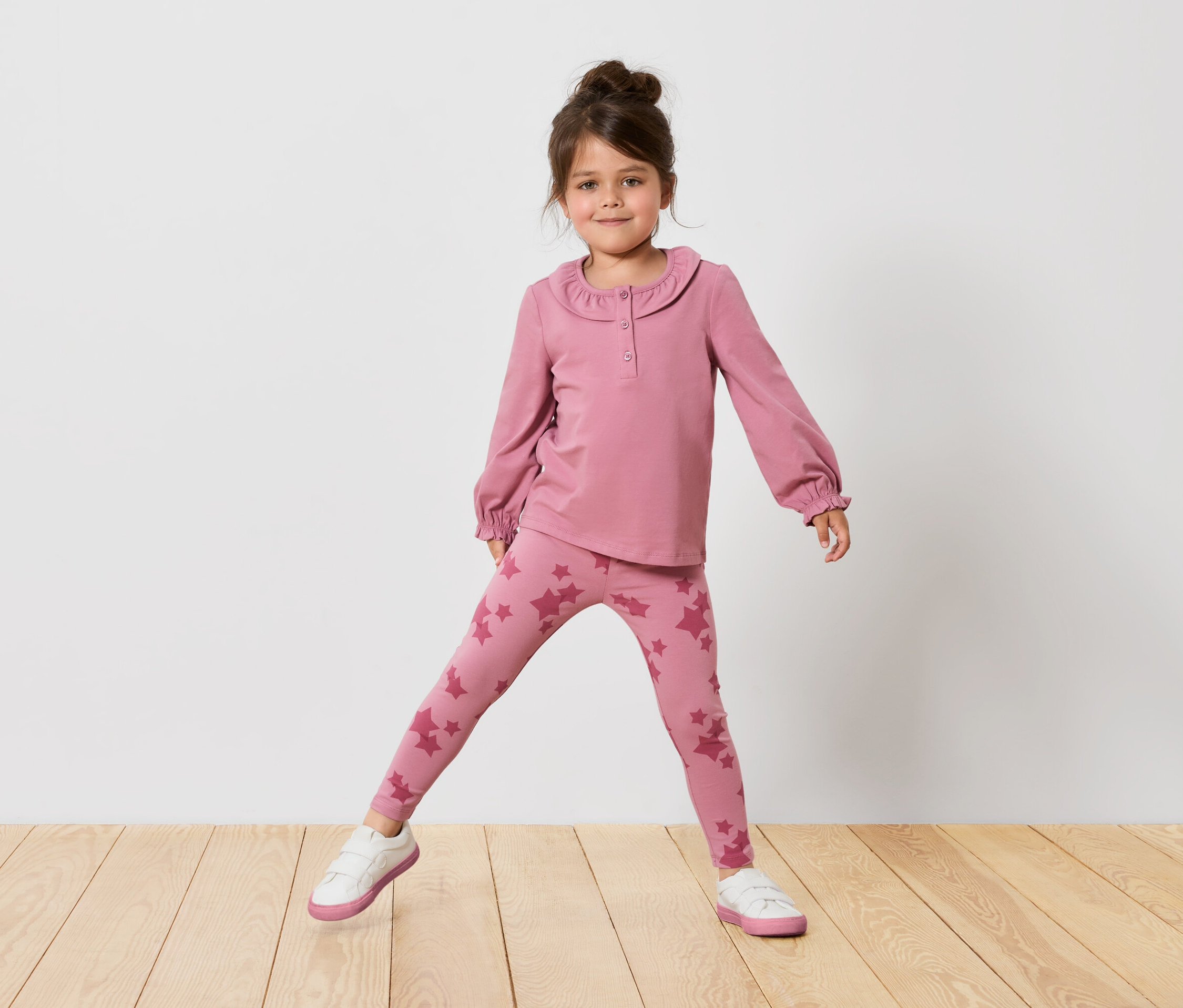 Dívka v růžovém topu s dlouhým rukávem a legínách s hvězdami stojí na dřevěné podlaze před bílou zdí.