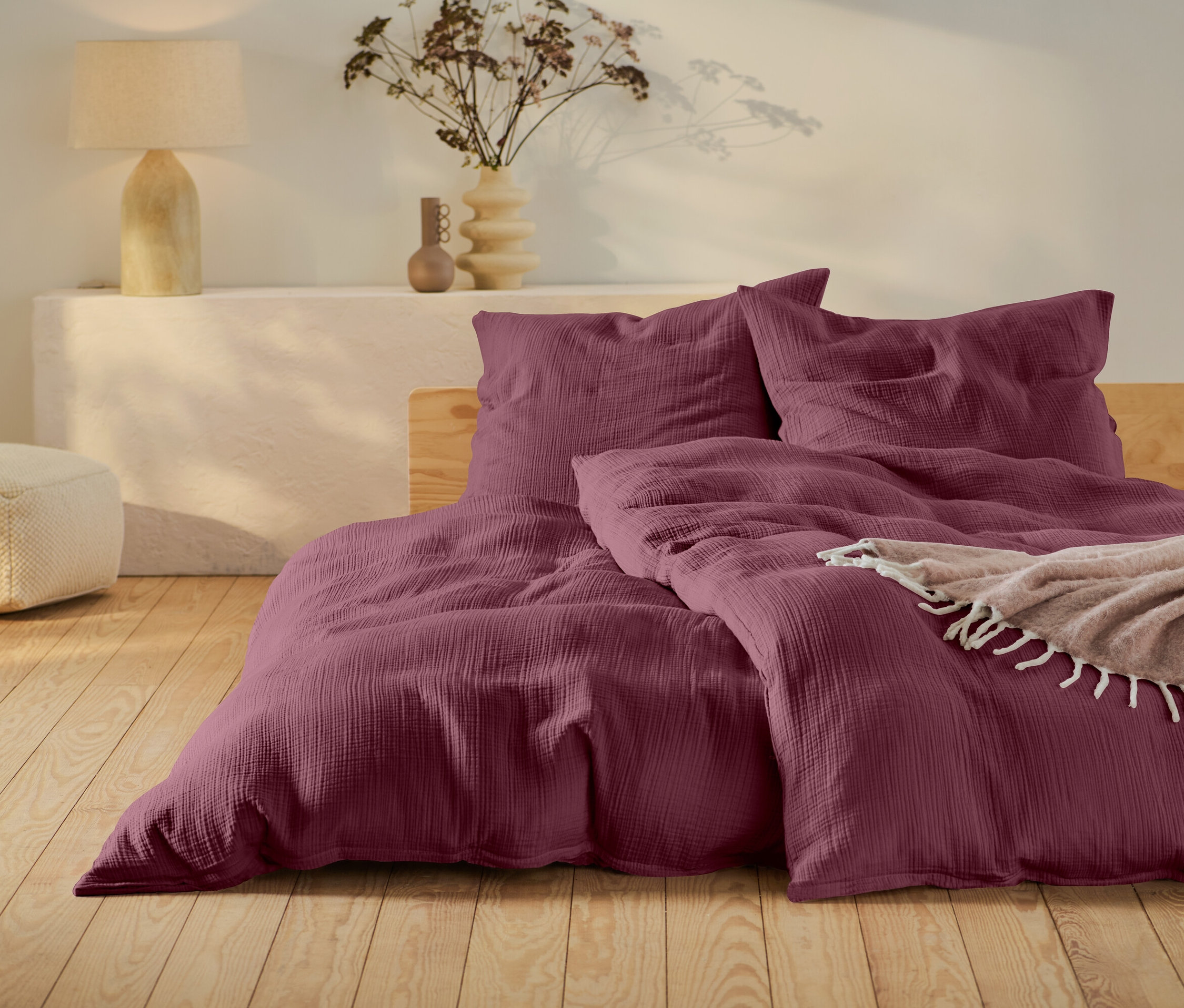 Vínové ložní prádlo na posteli s dřevěným rámem, béžový puf a lampa na bílém stolku.