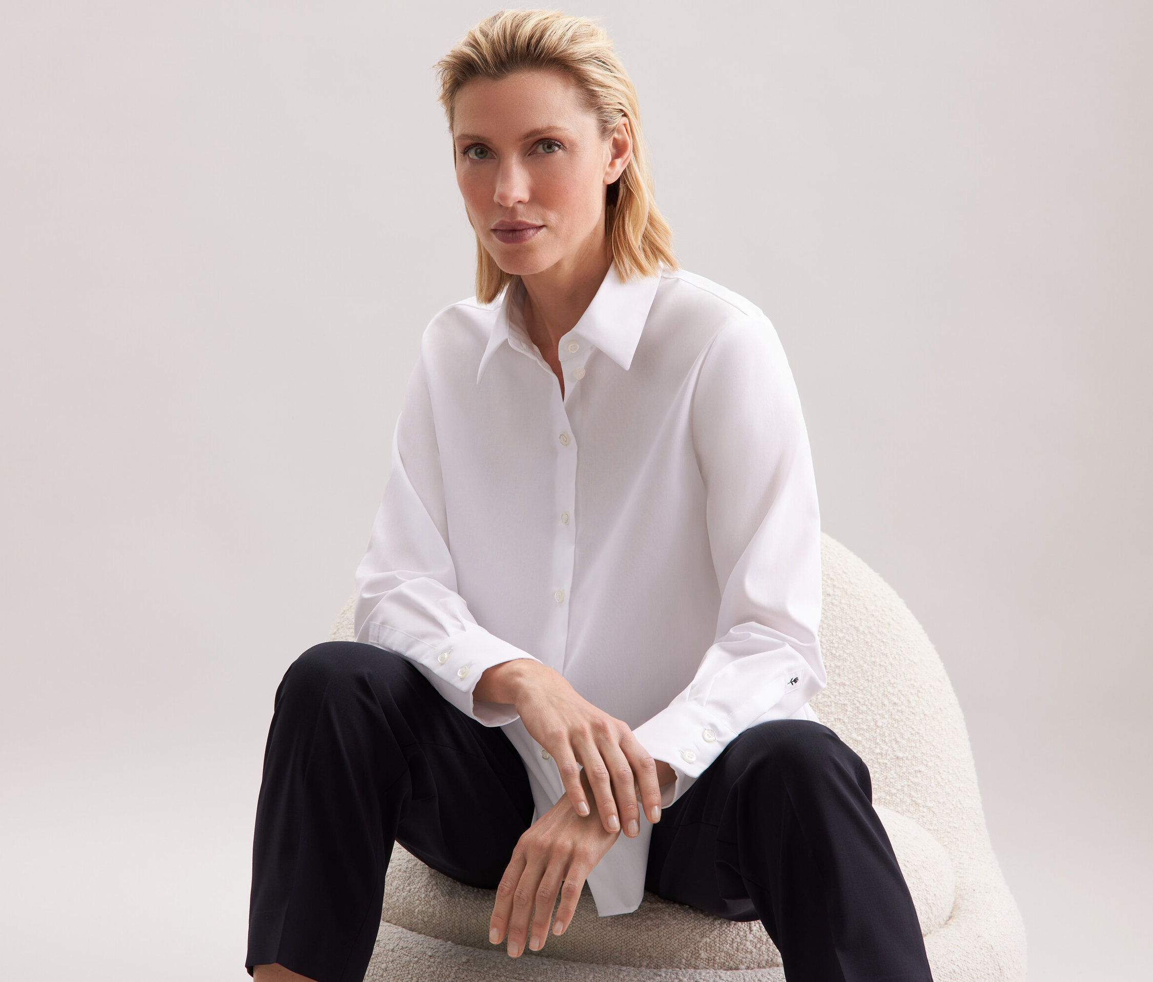 Žena sedí na židli a má na sobě bílou košili a černé kalhoty.