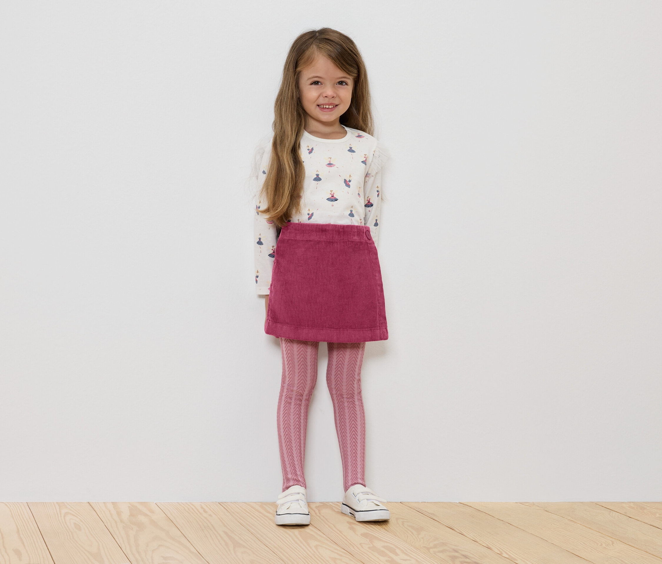 Dívka stojí a má na sobě bílé tričko s dlouhým rukávem s baletkami, růžovou manšestrovou sukni, růžové vzorované legíny a bílé tenisky.