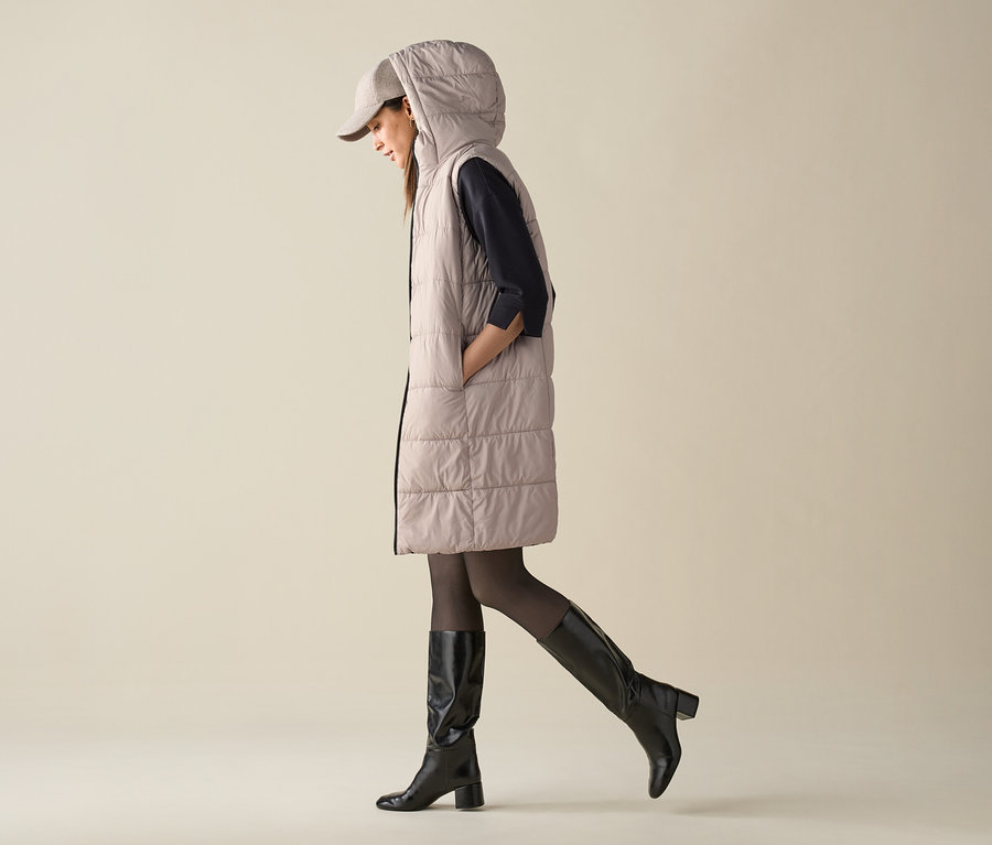 Žena kráčí a má na sobě prošívanou vestu s kapucí, čepici a kozačky.