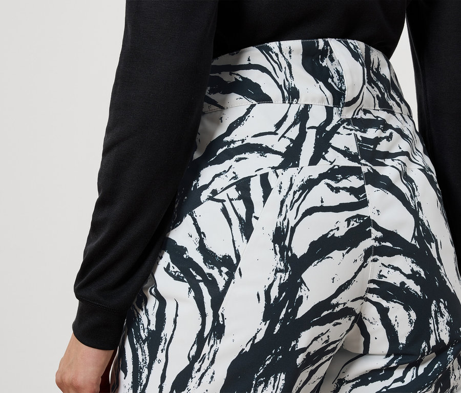 Pohled zezadu na ženu v černém topu a bílých kalhotách se vzorem černých větví.