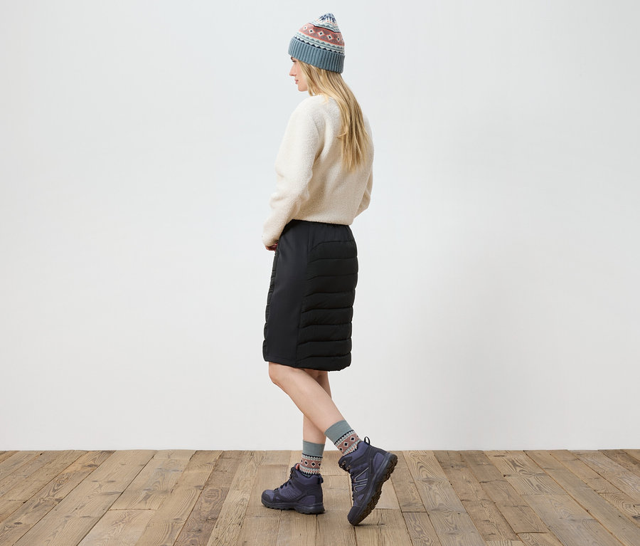 Žena pózuje v krémovém svetru, černé sukni, botách a pletené čepici.