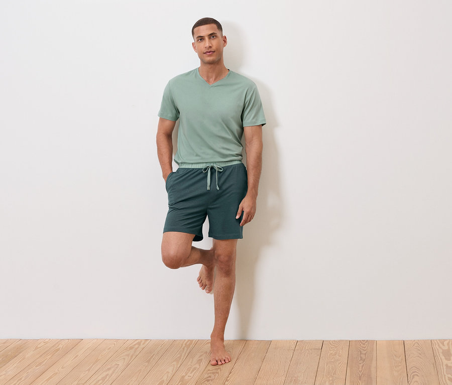 Muž stojí na jedné noze, opírá se o zeď, má na sobě zelené tričko a šortky.