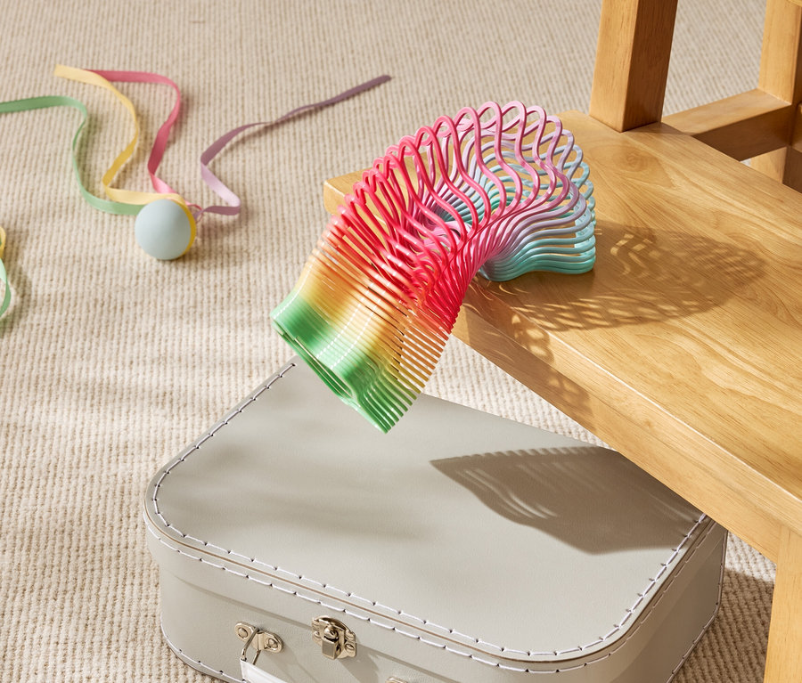 Barevná hračka spirála leží částečně na béžovém kufru pod dřevěnou židlí.