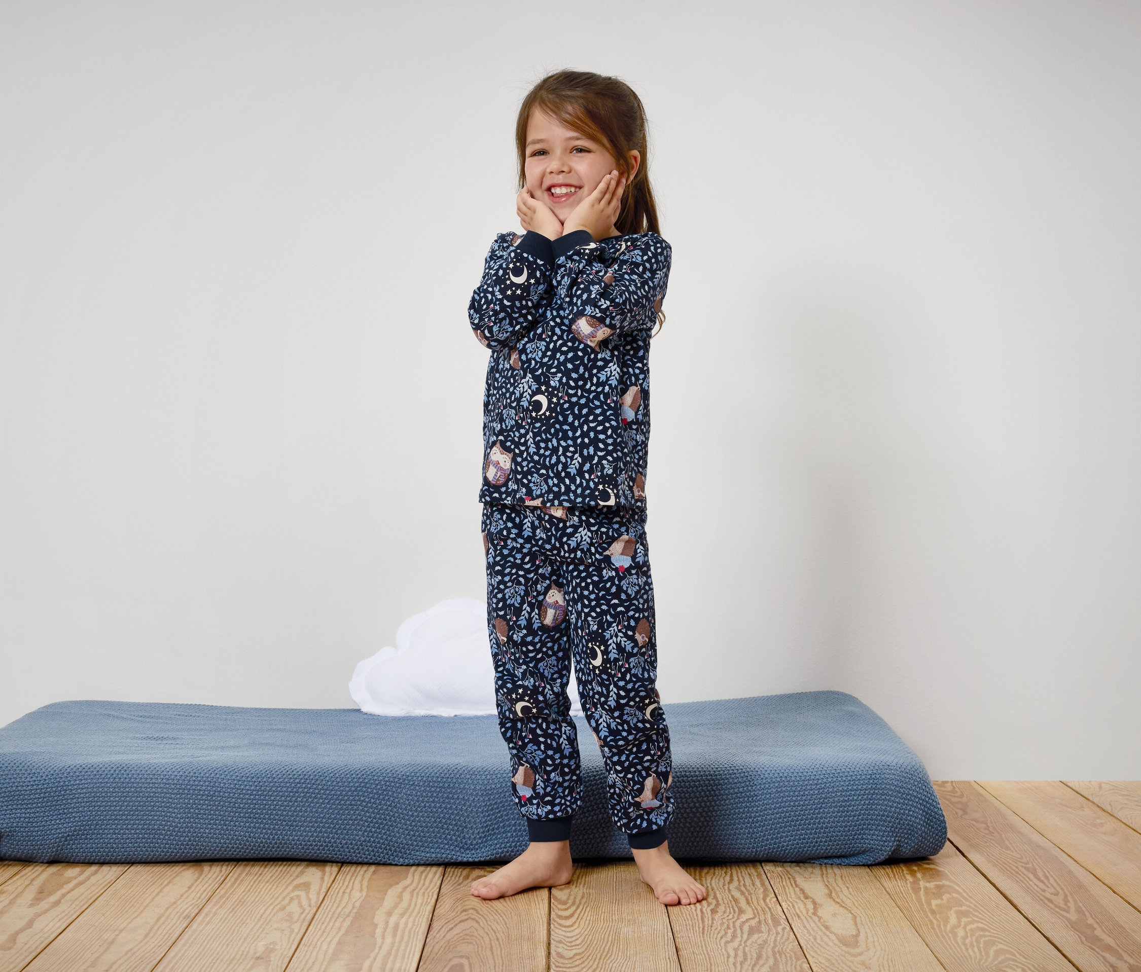 Usmívající se dívka v modrém pyžamu s potiskem stojí na dřevěné podlaze.