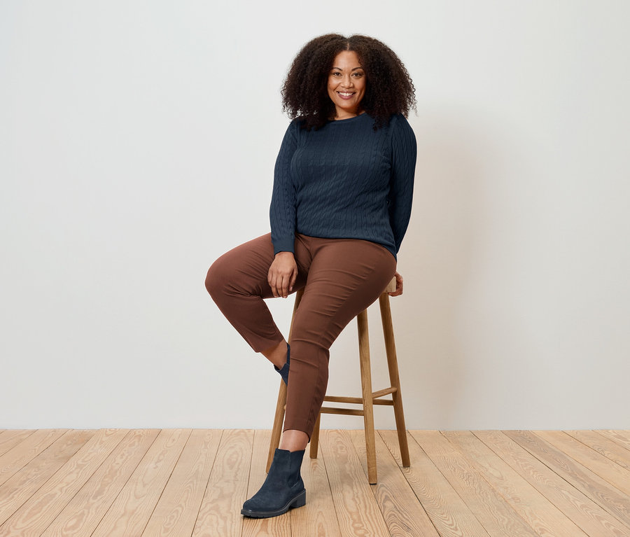 Žena sedí na dřevěné stoličce, má na sobě tmavě modrý svetr, hnědé kalhoty a modré boty.