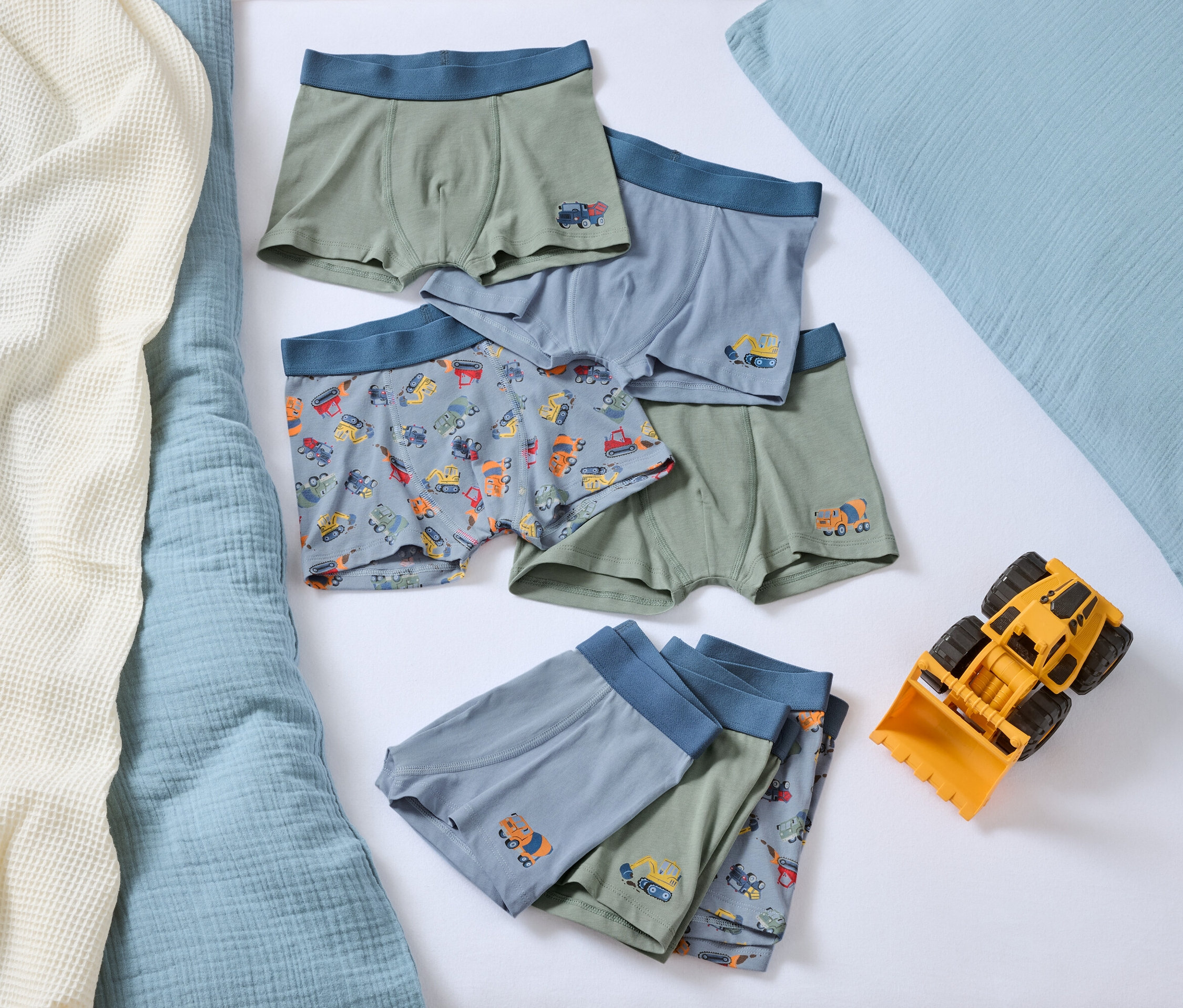 Na posteli leží hromada chlapeckých boxerek s motivy autíček a žlutá hračka buldozeru.